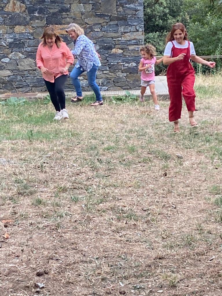 3 chicas y una niña juegan en el campo, es una escena de película, el vestuario es contemporaneo y colorido, un mono rojo, camisa naranja, camisa azul, camiseta rosa. La imagen es alegre y divertida