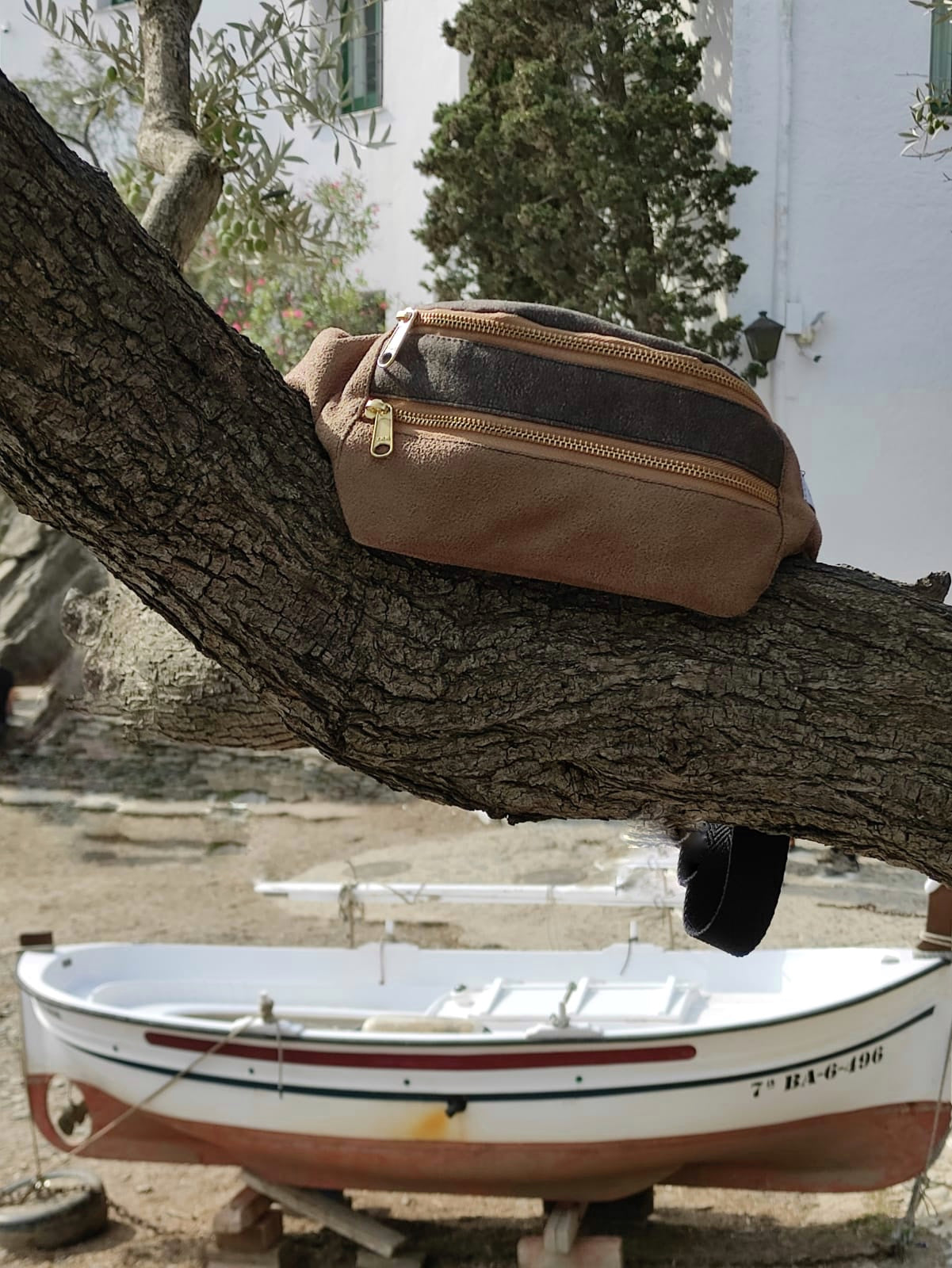 Foto de la riñonera sobre el tronco de un árbol, con fondo marítimo, un barco de pescadores que evoca un viaje.